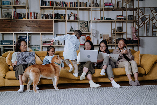 Family of six sitting on a sofa with a dog, while members read, use a laptop, and a smartphone.